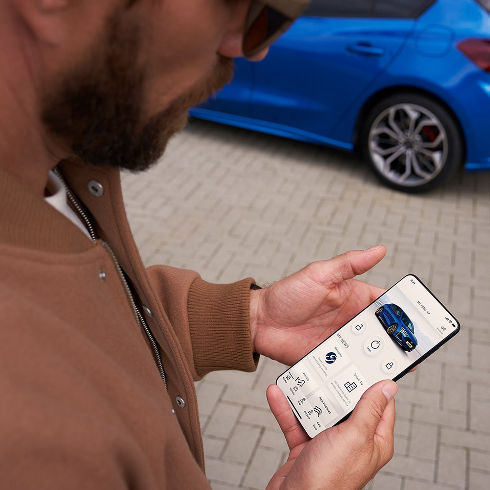 Une personne tient un smartphone sur lequel on voit l’application FordPass™. Une Ford Focus couleur bleue se trouve à l’arrière-plan.