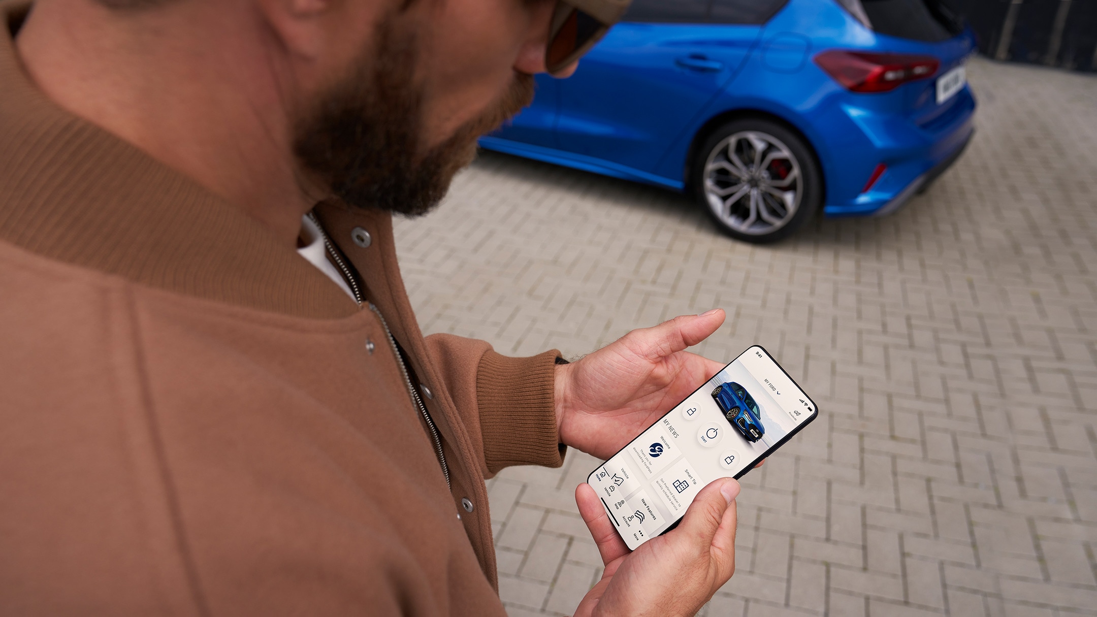 L’application FordPass. Un homme regarde son smartphone. Une Ford Focus couleur bleue en arrière-plan.