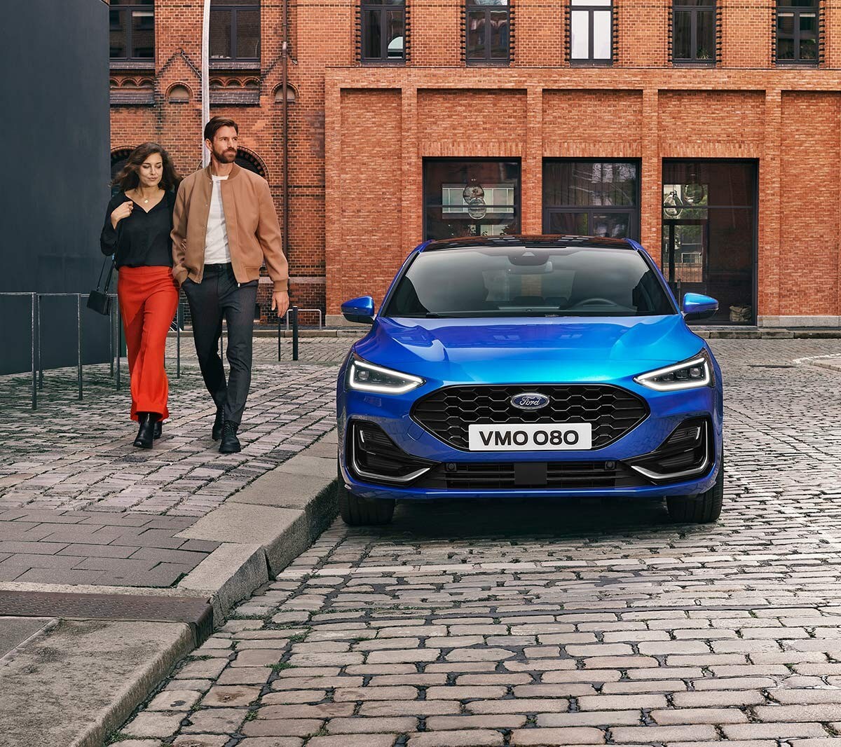 Ford Focus couleur bleue. Vue de face, se garant dans une zone industrielle. Deux personnes passent à côté.
