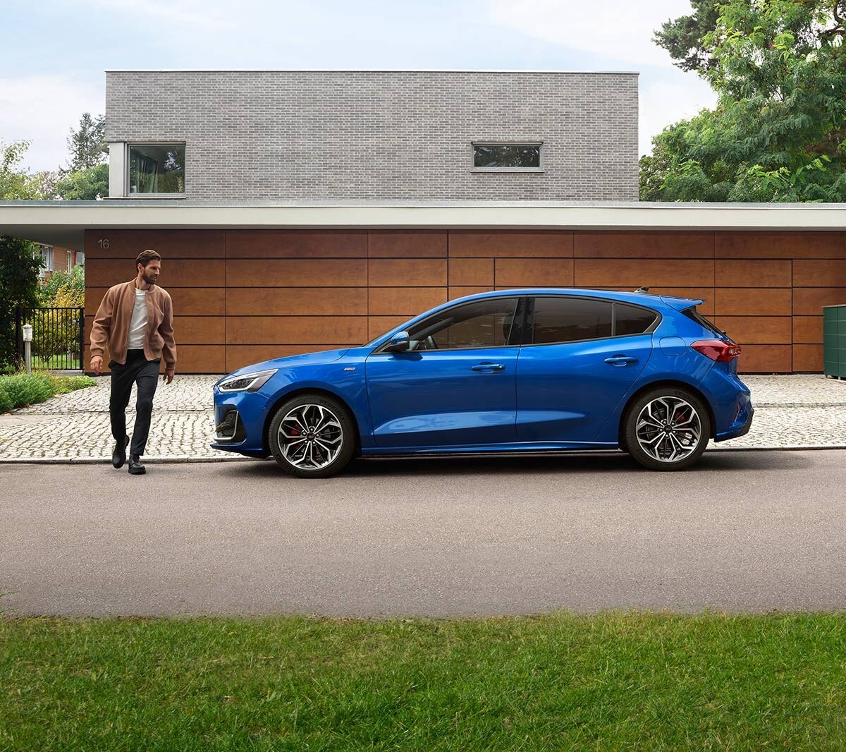 Ford Focus couleur bleue. Vue latérale, garée devant une maison moderne. Un homme court à côté.