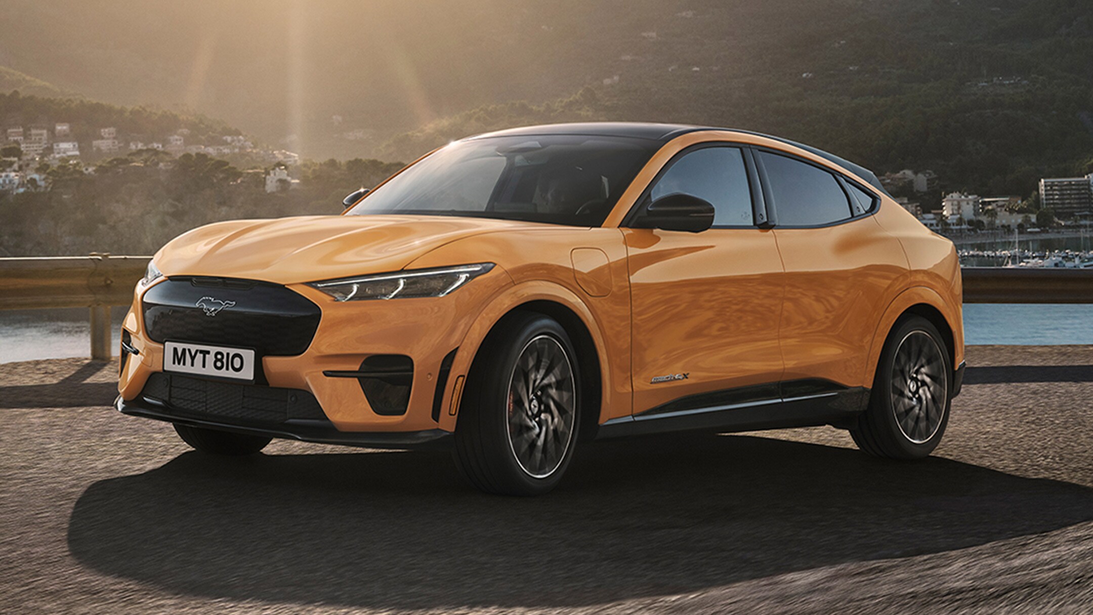 Ford Mustang Mach-E GT couleur orange. Vue de face aux trois quarts, roulant sur une route au coucher du soleil avec des montagnes en arrière-plan.