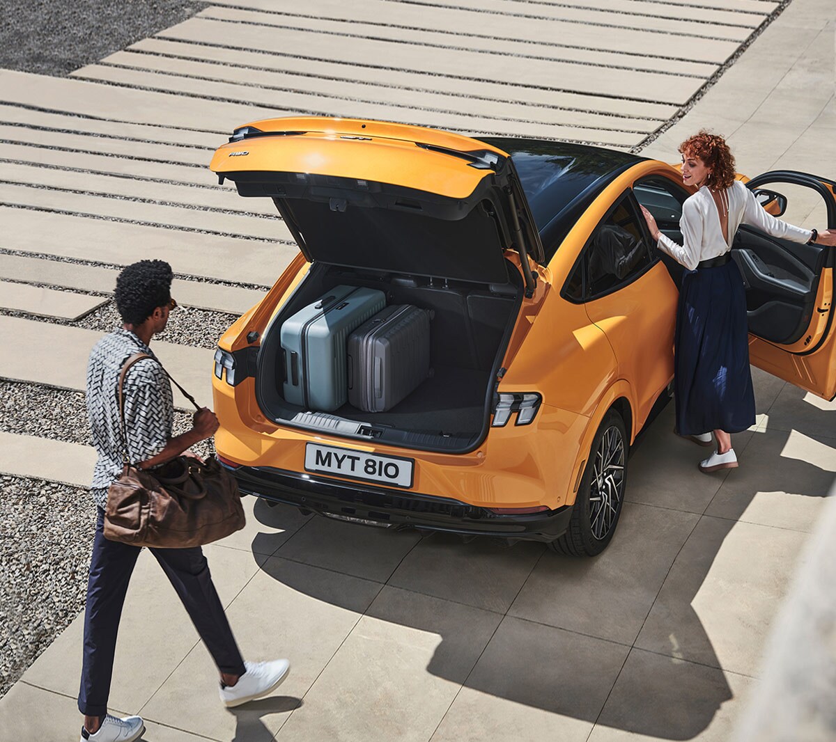 Ford Mustang Mach-E GT en orange. Vue de trois-quarts arrière, deux personnes utilisent le hayon commandé par capteur.