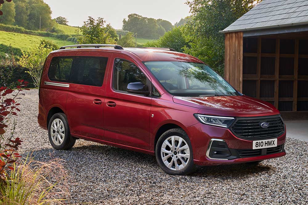 Ford Grand Tourneo Connect couleur rouge, vue de face aux trois quarts, stationnant sur un chemin de gravier, devant une habitation