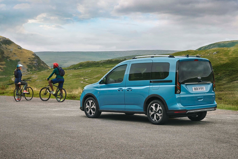 Un Ford Tourneo Connect bleu est garé sur une route dans un paysage verdoyant et vallonné, avec deux personnes à vélo à proximité.