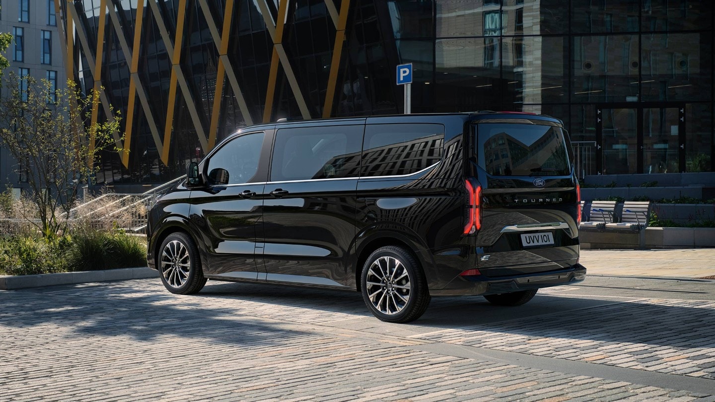 Ford Tourneo Custom couleur noire, vue arrière aux trois quarts, garé sur une place de parc devant des bâtiments modernes