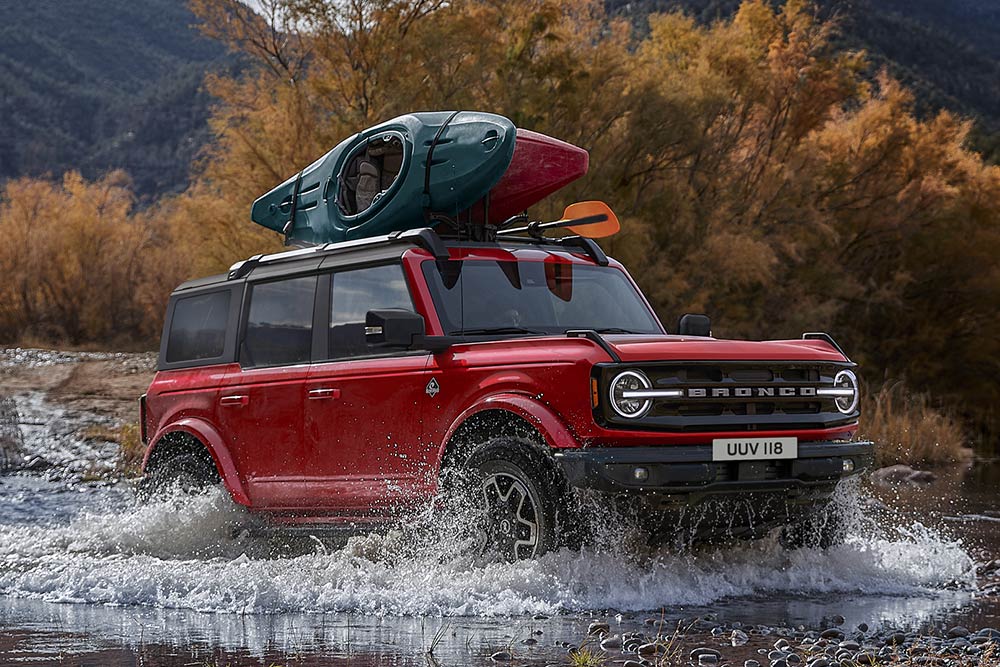Vue latérale d'un Ford Bronco rouge traversant des eaux peu profondes dans un paysage de montagne.