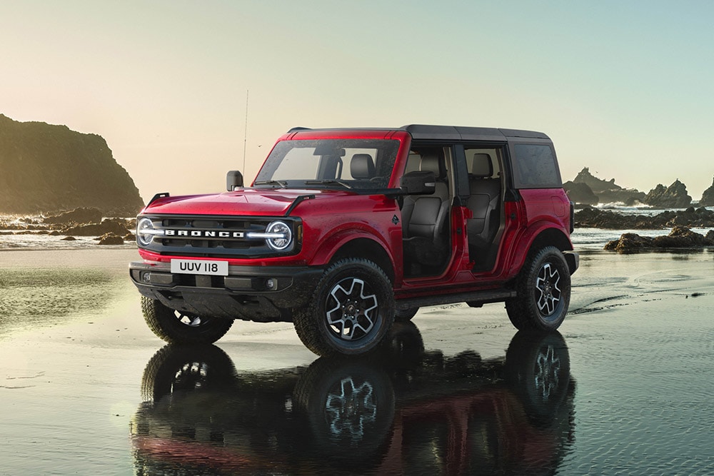 Vue latérale d'un Ford Bronco rouge sans portes, stationné sur eine plage de sable mouillé.