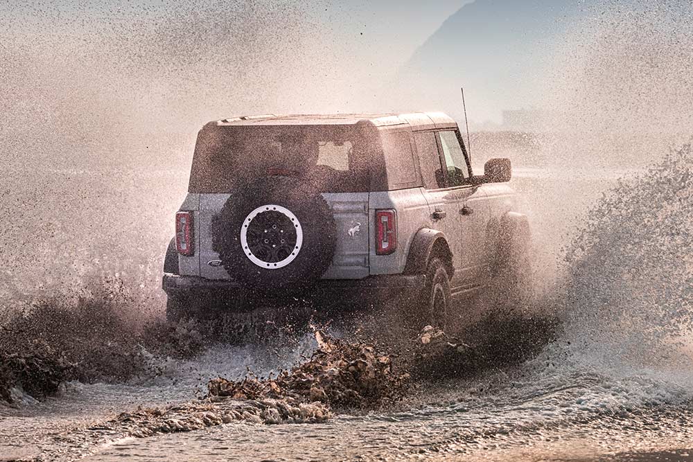Vue arrière d'un Ford Bronco argenté traversant des gerbes d'eau sur la plage.