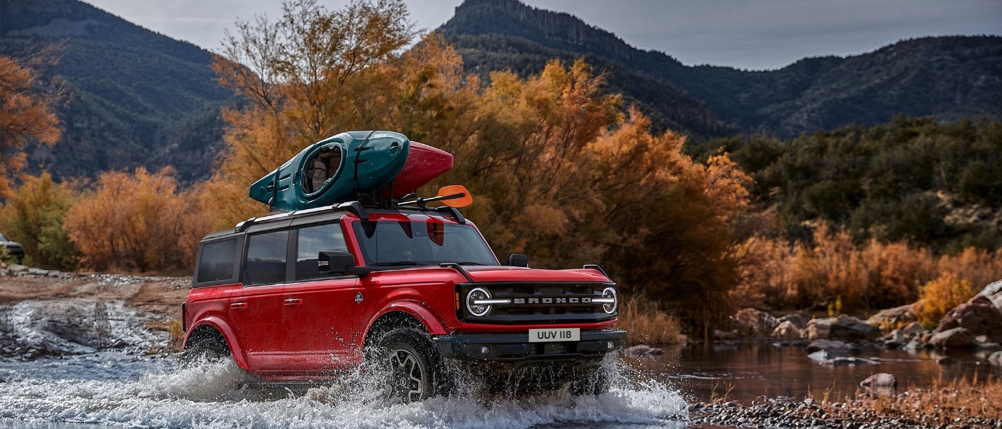 Ford Bronco couleur rouge. Vue de face aux trois quarts, traversant une rivière dans les montagnes.