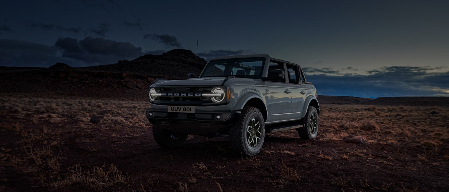 Ford Bronco couleur grise. Vue de face aux trois quarts, roulant dans la nature au coucher du soleil.