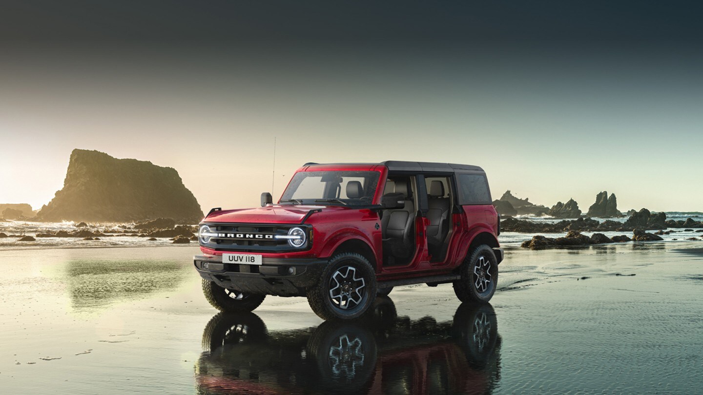 Ford Bronco couleur rouge. Vue de face aux trois quarts, traversant une rivière dans les montagnes