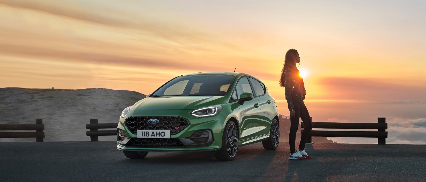 Ford Fiesta ST couleur verte. Vue avant au trois quarts, une femme admire le soleil couchant.