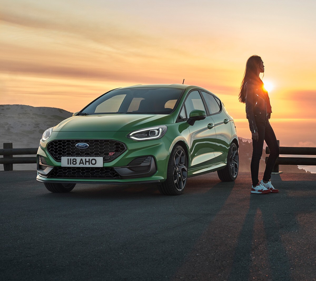 Ford Fiesta ST couleur verte. Vue de face aux trois quarts, femme admirant le soleil couchant.