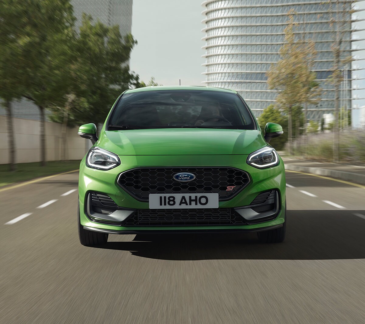 Ford Fiesta ST couleur verte. Vue de face, roulant sur une route devant des arbres et un bâtiment moderne.