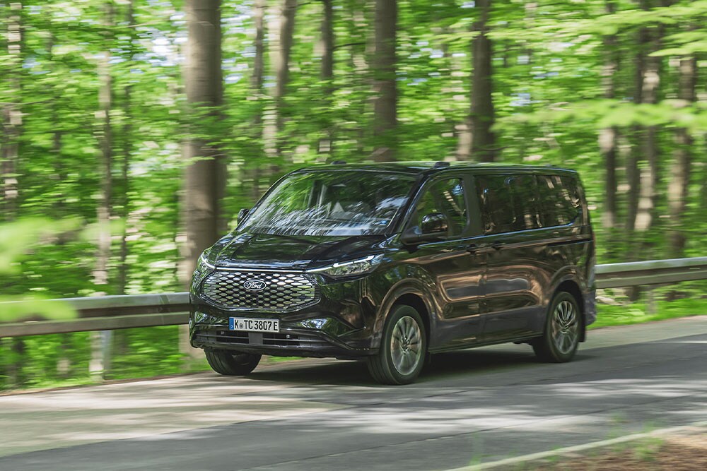 Ford E-Tourneo Custom noir roulant sur une route bordée d'arbres.