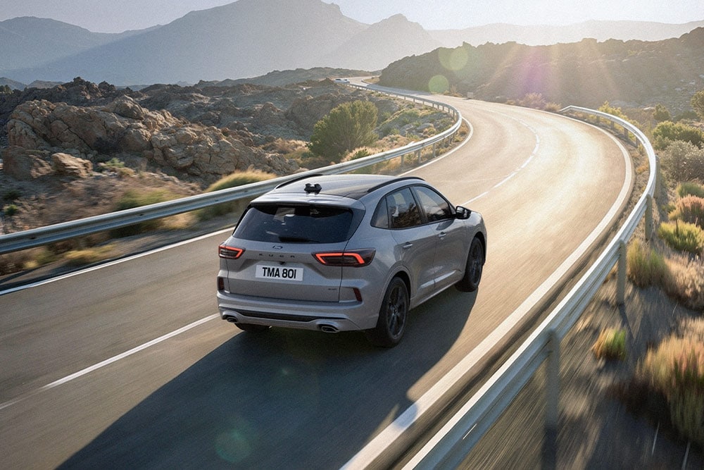 Ford Kuga gris roulant sur une route pittoresque, avec un paysage de montagne ensoleillé en arrière-plan.