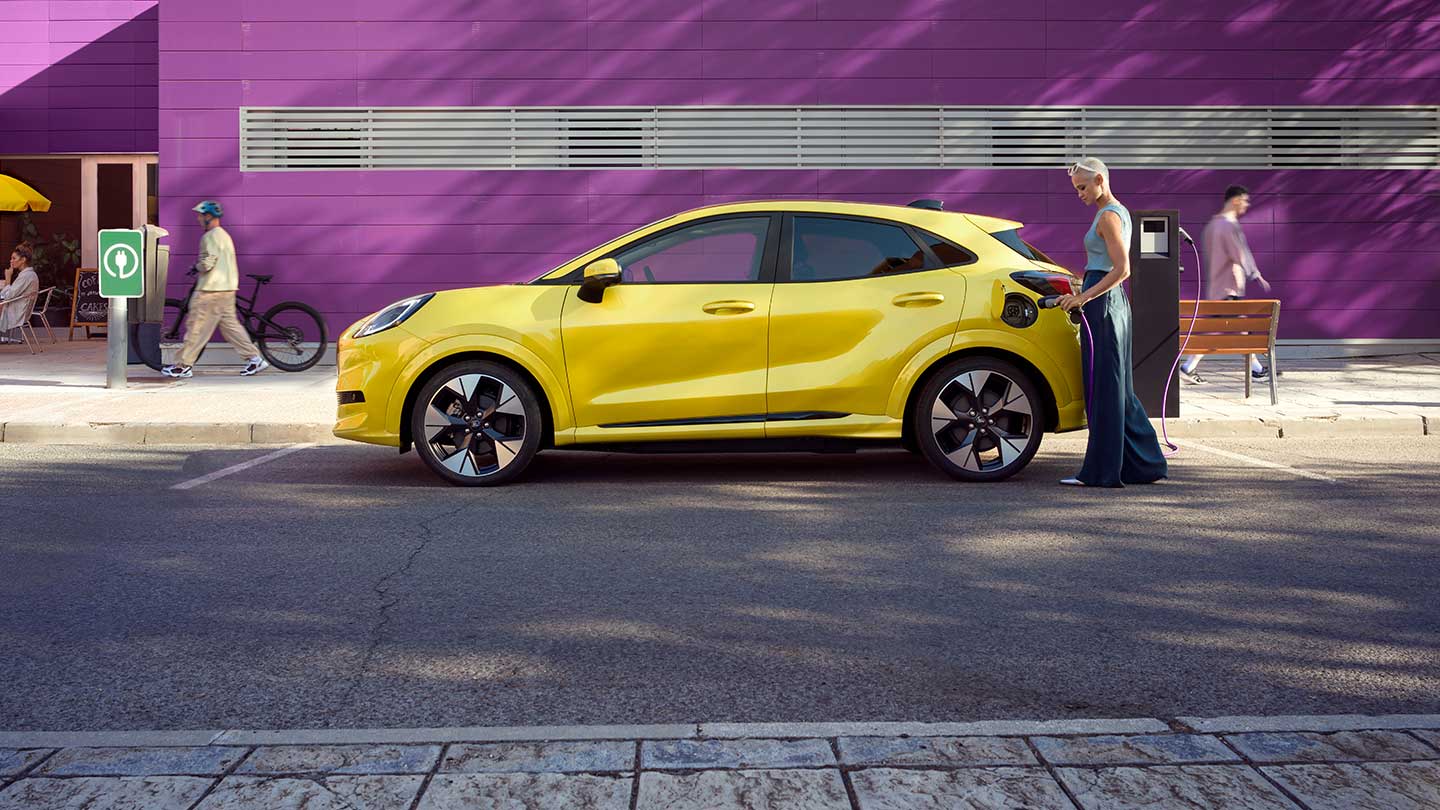 Vue latérale d’une Ford Puma Gen-E® électrique jaune branchée à une station de recharge publique sur une place de parking.