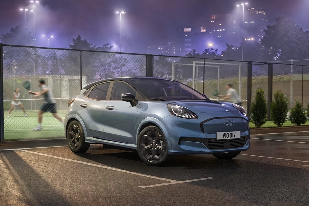 Un Ford Puma Gen-E bleu est garé à côté d’un court de tennis, avec les lumières de la ville en arrière-plan.