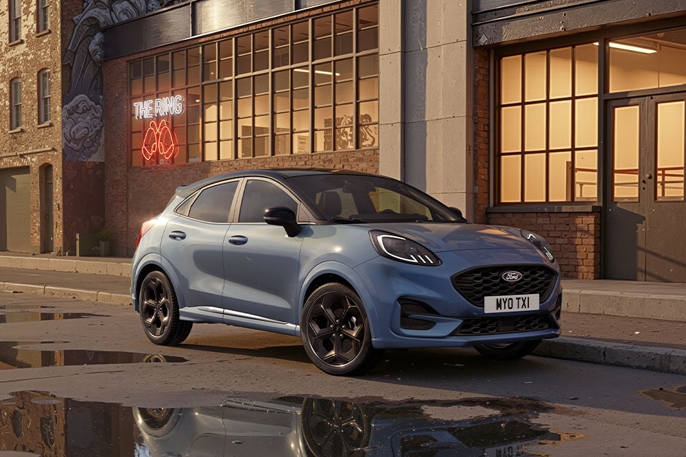 Un Ford Puma bleu est garé dans une rue de la ville au coucher du soleil, des flaques d’eau reflètent les bâtiments.