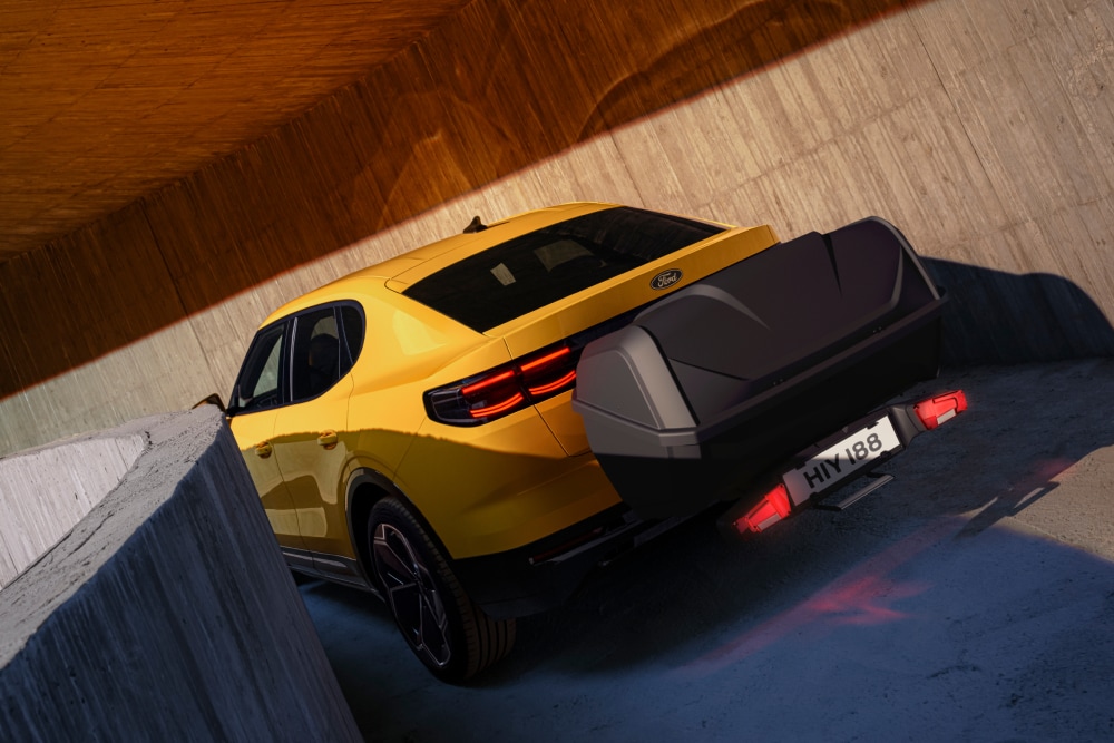 Vue arrière d’une Ford Capri jaune, stationnée dans un bâtiment moderne en béton, avec un coffre à bagages monté sur l’attelage.