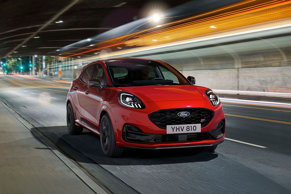 Un SUV de sport Ford Puma ST rouge roulant de nuit sur une route urbaine fortement éclairée.