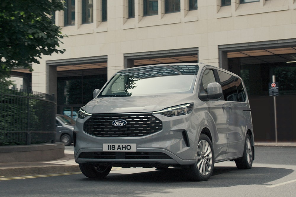 Vue avant trois quarts du Tourneo Custom gris roulant près d’un immeuble en béton.