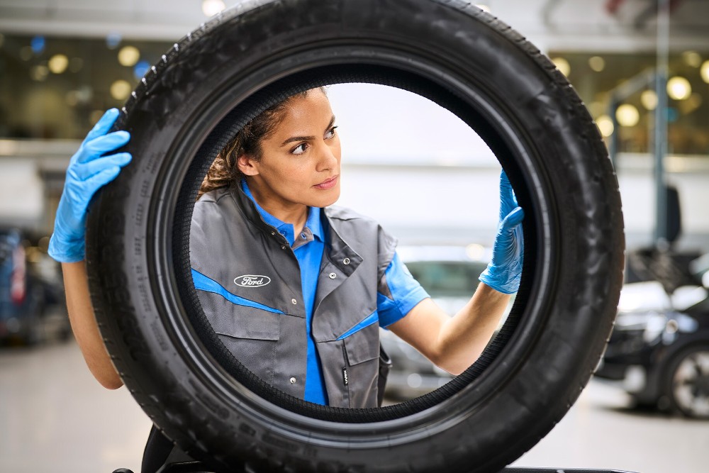 Un technicien Ford en gants bleus inspecte un pneu tenu en l'air dans un atelier.