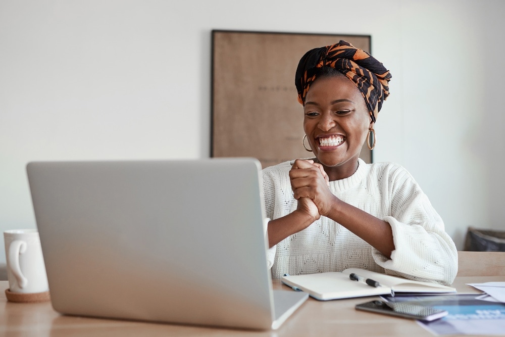 A person with a happy expression looks at a laptop, hands clasped in excitement at a desk.