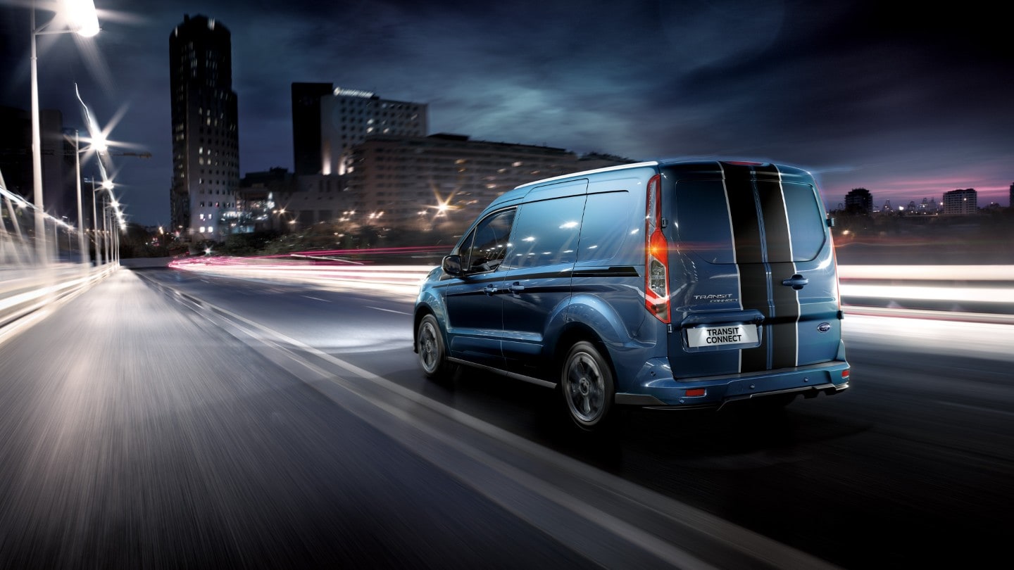 Ford Transit Connect couleur bleue, vue arrière aux trois quarts traversant une grande ville de nuit