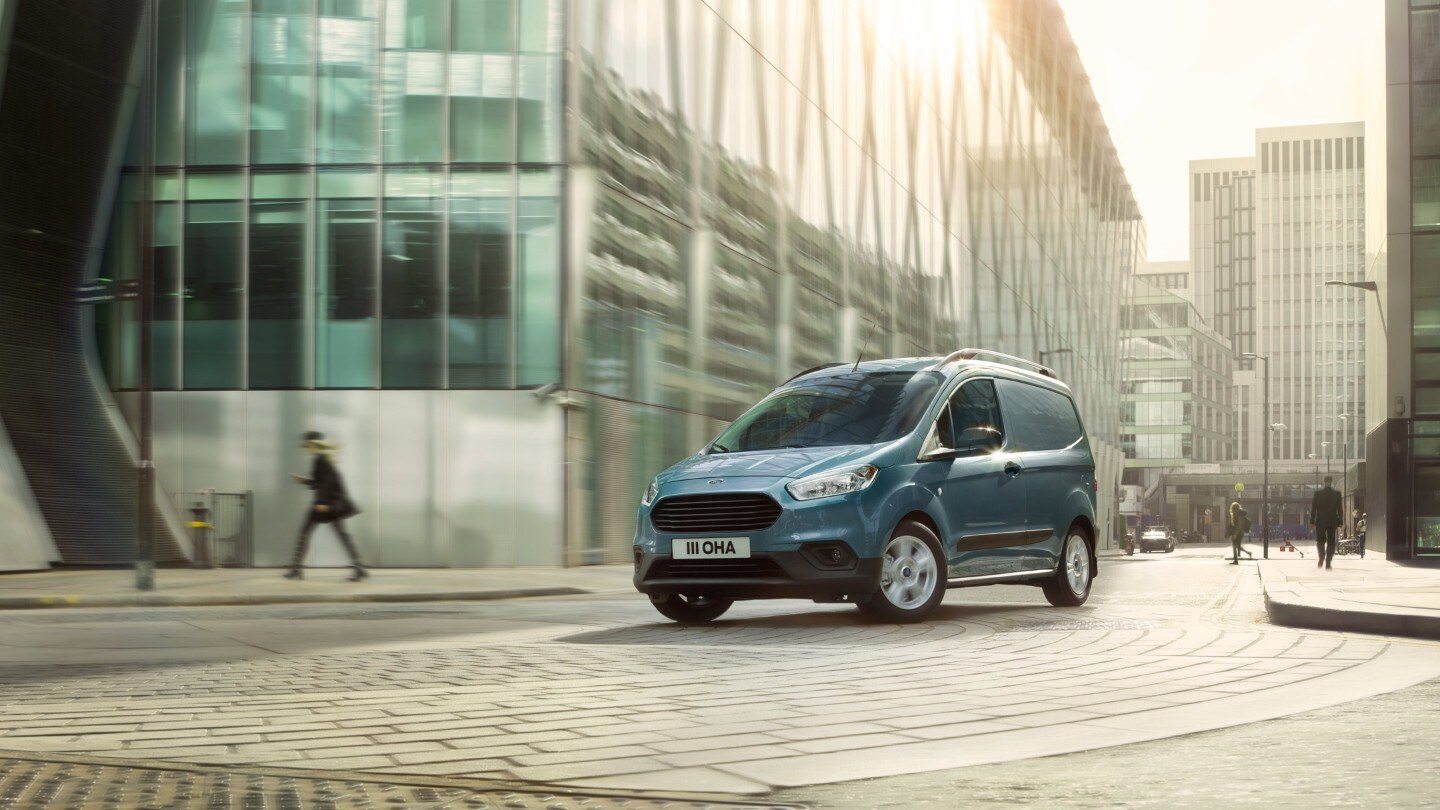 Ford Transit Courier couleur grise, vue de face aux trois quarts, roulant dans des rues étroites d’un centre-ville