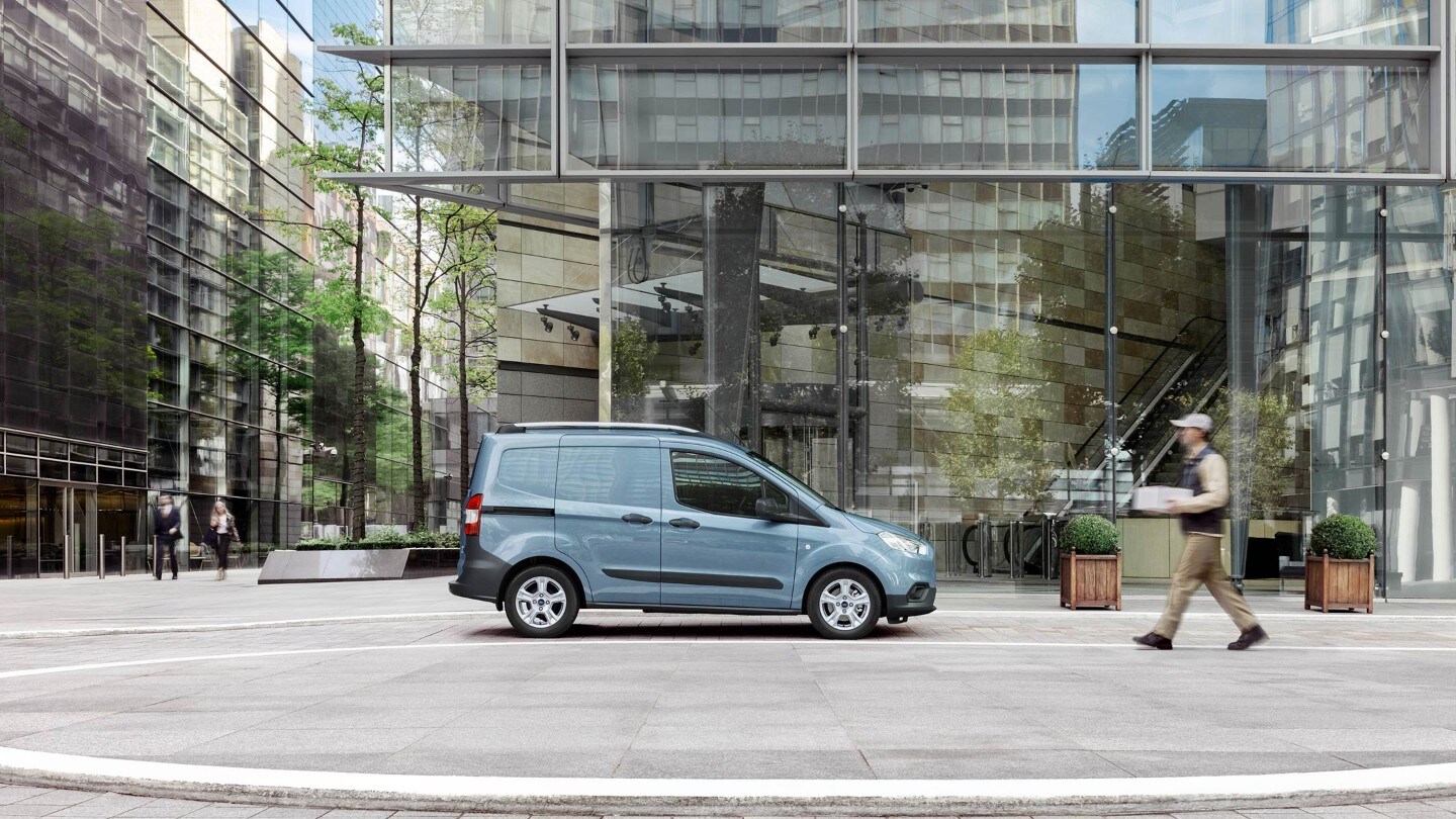 Ford Transit Courier couleur grise, vue latérale, stationnant devant un bâtiment moderne avec façade en verre