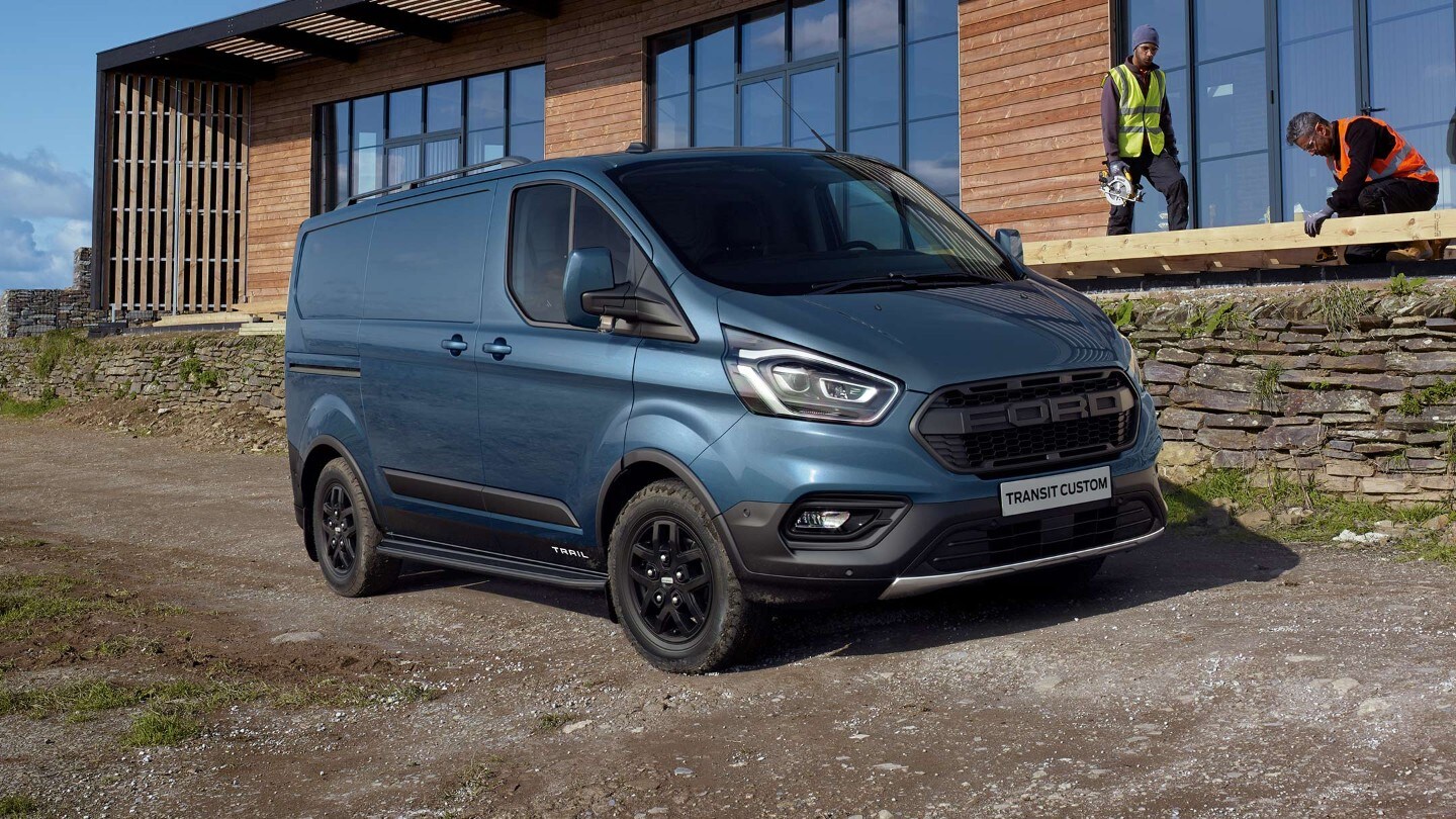Fourgon Ford Transit Custom Trail bleu Vue ¾ latérale. Stationnement sur une route de chantier, devant un bâtiment 
