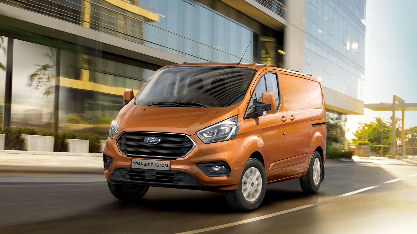 Ford Transit Custom couleur orange, vue de face aux trois quarts roulant devant un bâtiment avec une façade en verre