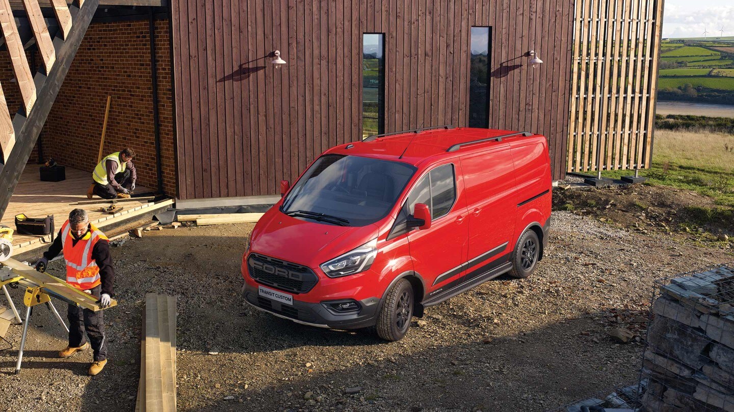 Ford Transit Custom Trail rouge, vue latérale aux trois quarts, parcs sur le chantier devant la maison en bois