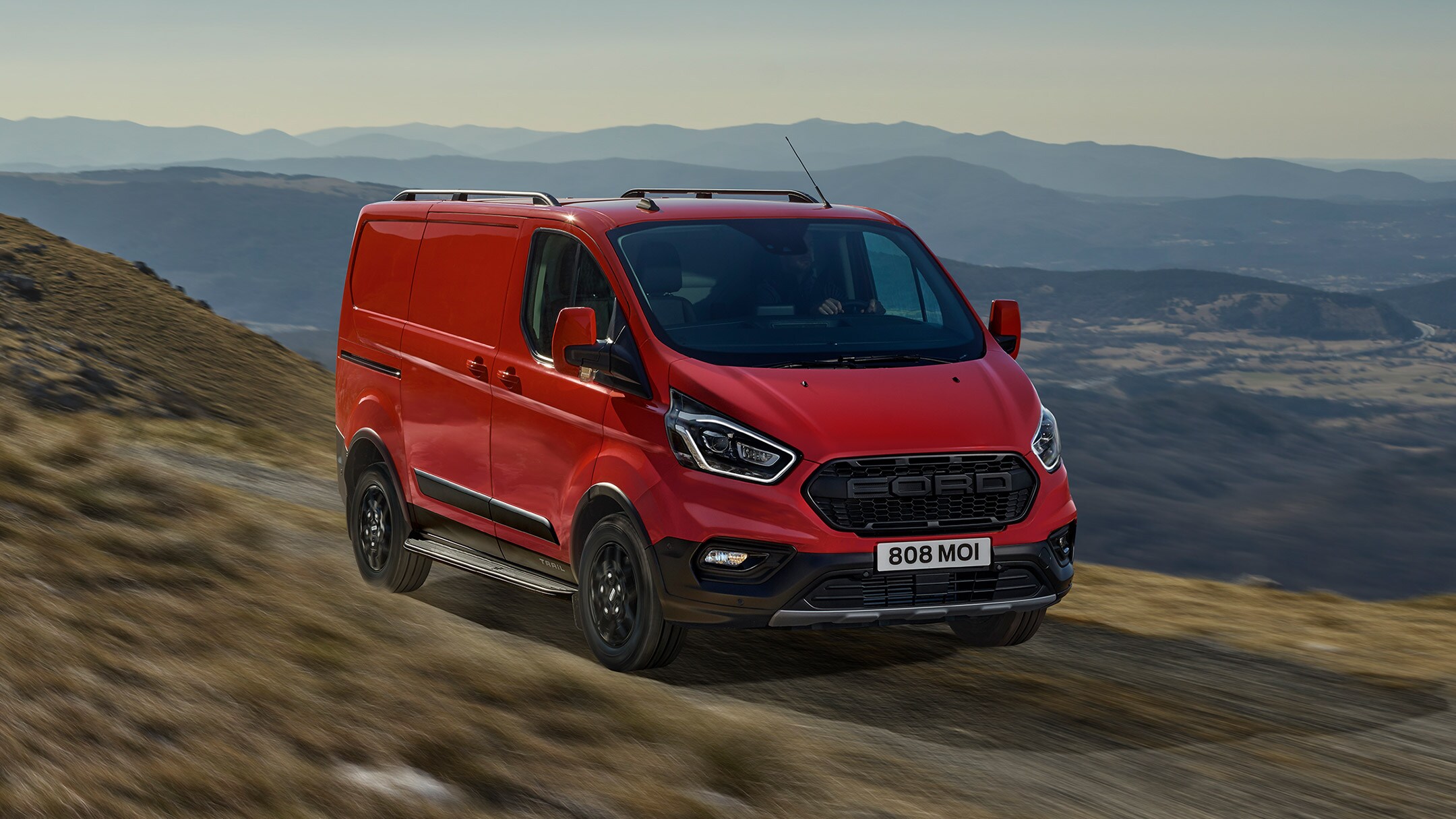 Fourgon Ford Transit Custom Trail rouge. Vue ¾ avant. Conduite sur un chemin de terre en zone montagneuse
