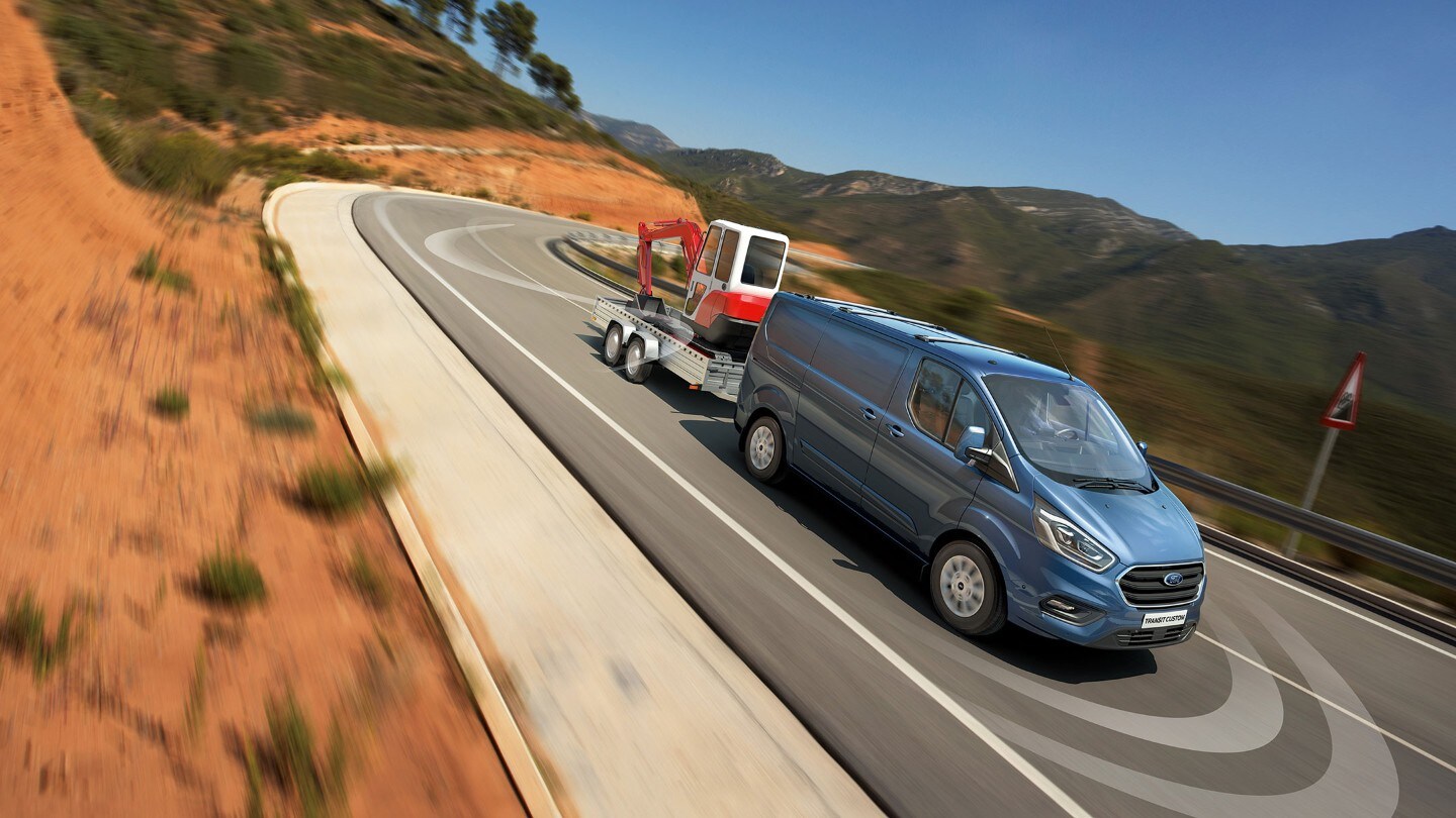 Ford Transit Custom couleur bleue, vue de face aux trois quarts avec remorque, descendant une route de campagne