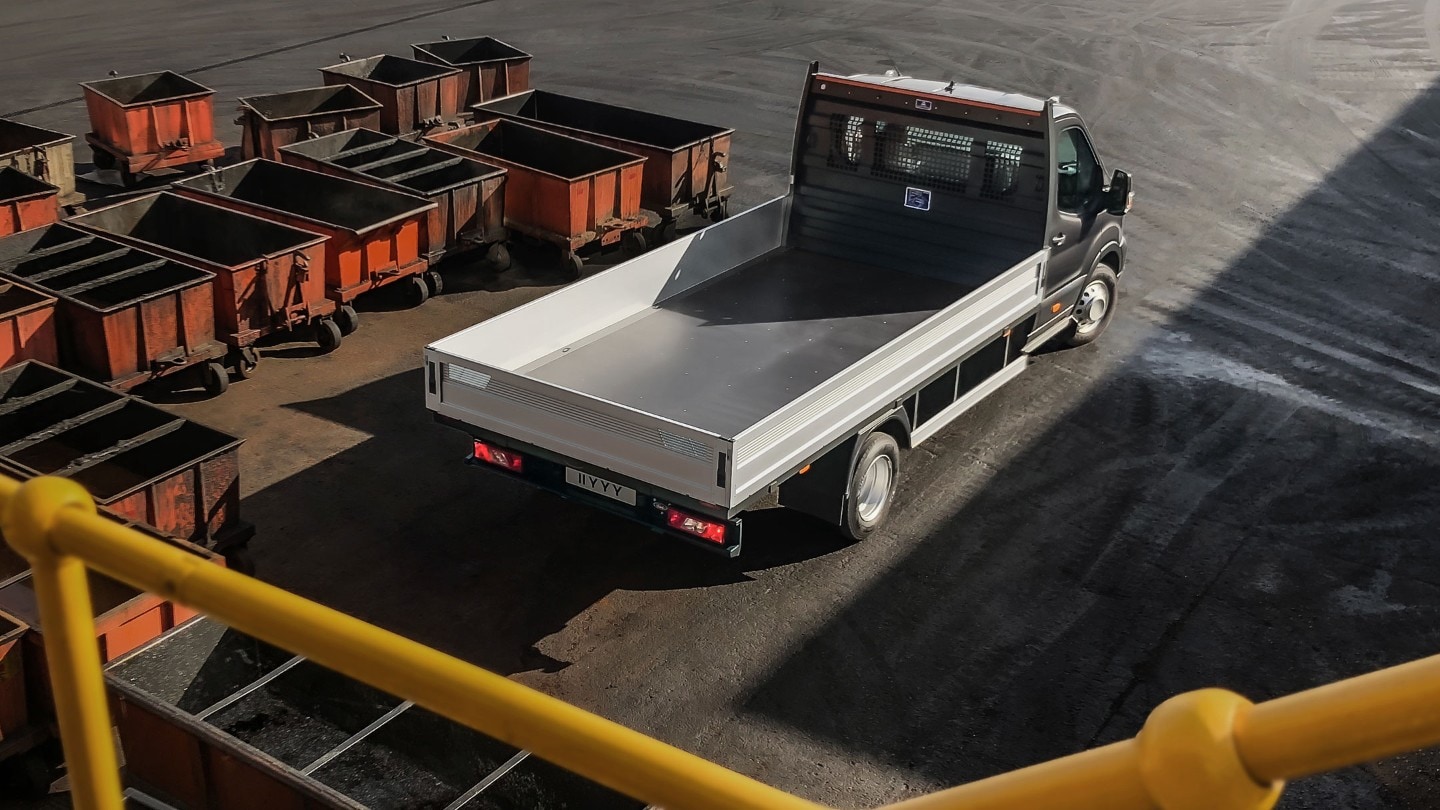 Vue aérienne arrière aux trois quarts sur un Ford Transit Chassis Cab 5 tonnes noir, garé sur une aire d’entreprise à côté de palettes chargées