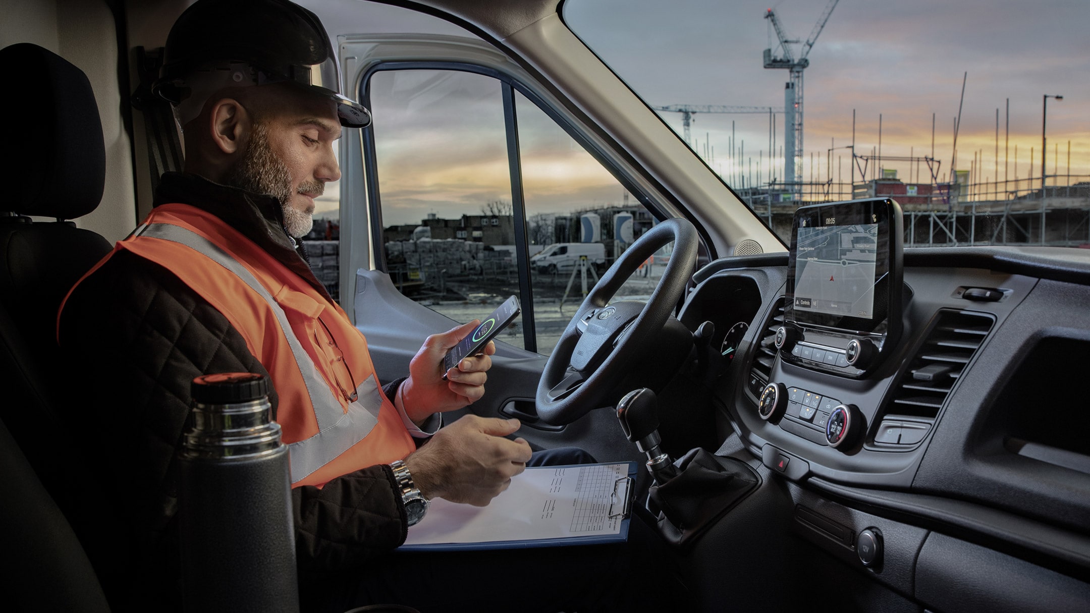 Ouvrier portant un casque de chantier assis derrière le volant d’un véhicule et regardant un téléphone portable