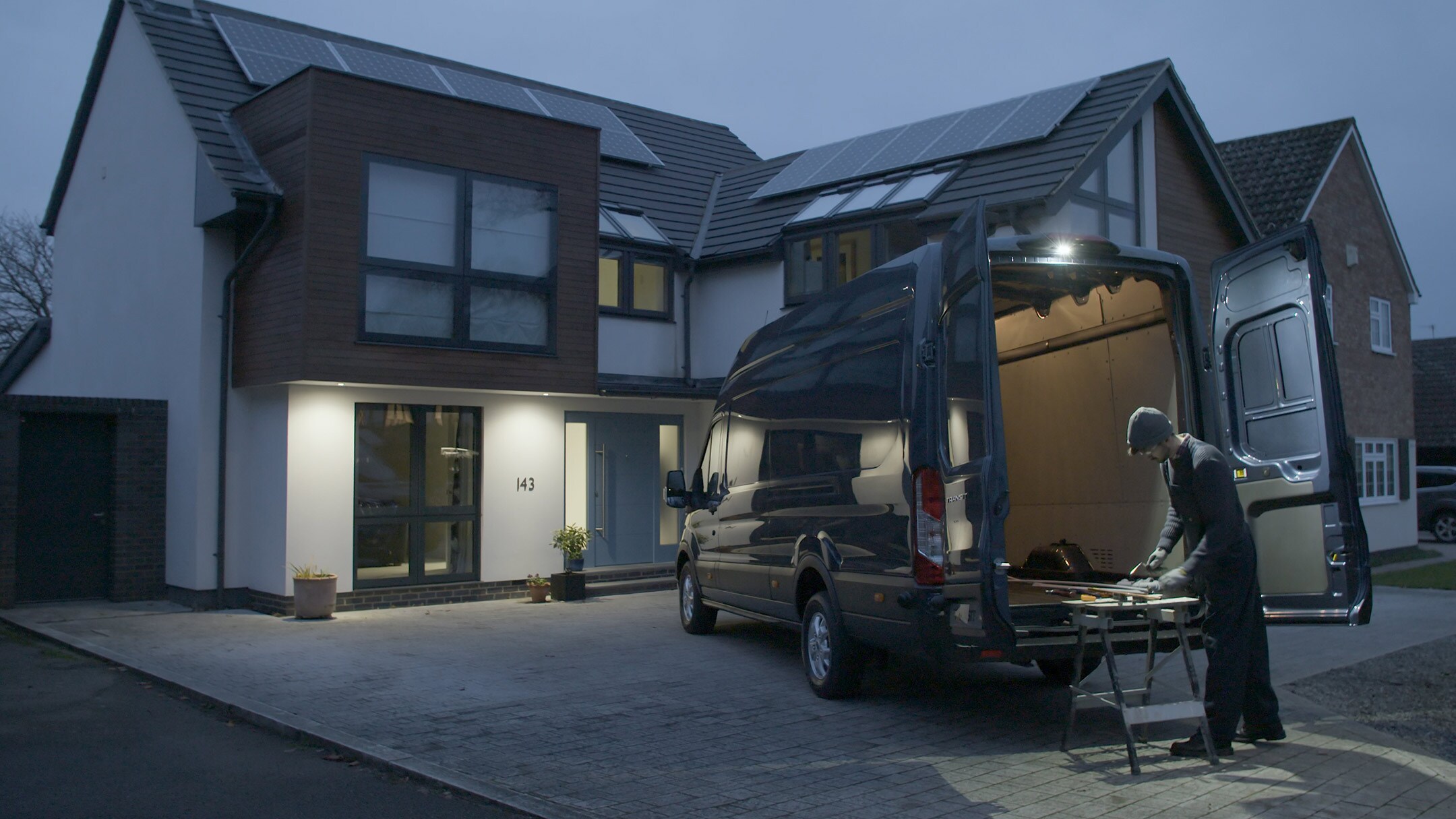 Ford Transit Fourgon couleur bleue, vue arrière aux trois quarts, portes arrière ouvertes, garé devant une maison dans l’obscurité avec un artisan.