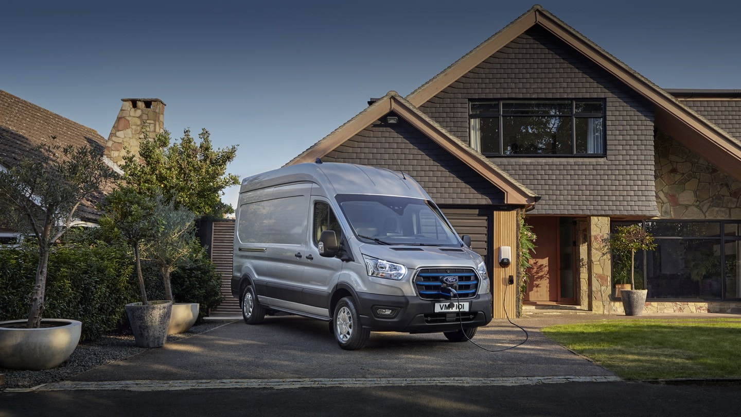 Ford E-Transit couleur argent, vue de face aux trois quarts, garé devant une maison et en cours de charge