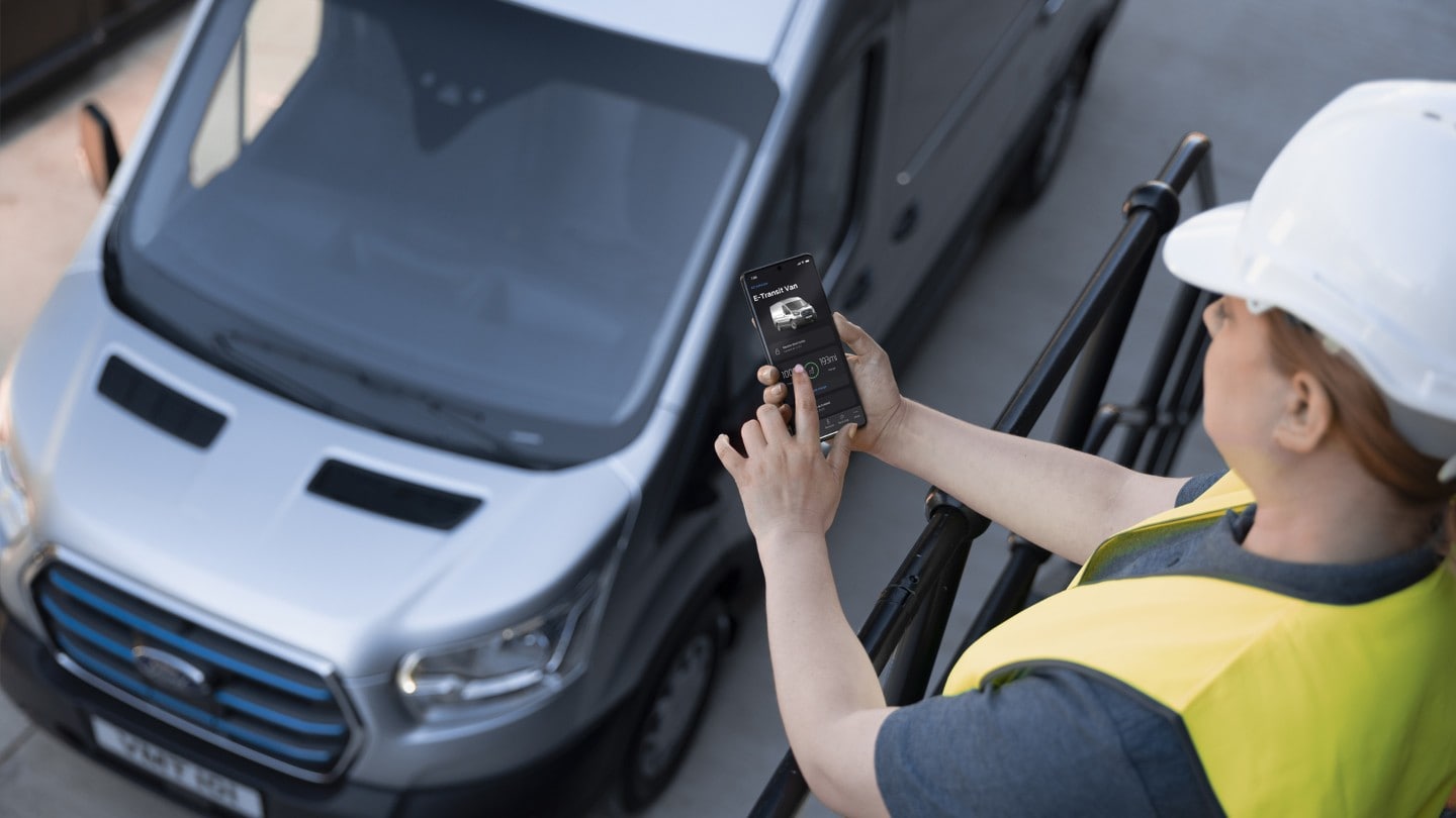 Femme avec casque de protection se tenant à côté du Ford E-Transit, un téléphone portable à la main