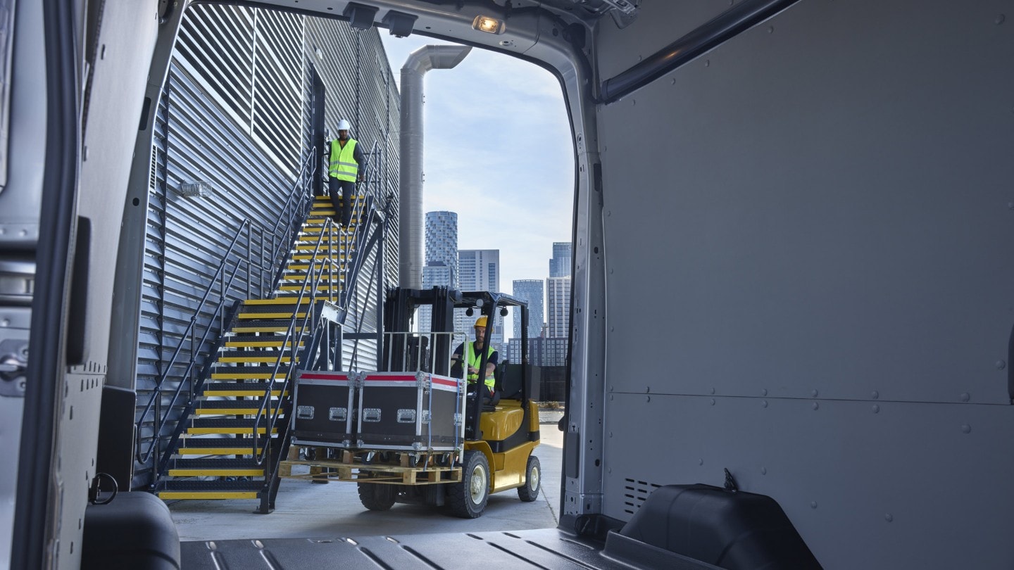 Ford E-Transit Fourgon, vue à travers la porte arrière ouverte depuis l’espace de chargement sur un chariot élévateur chargé en arrière-plan