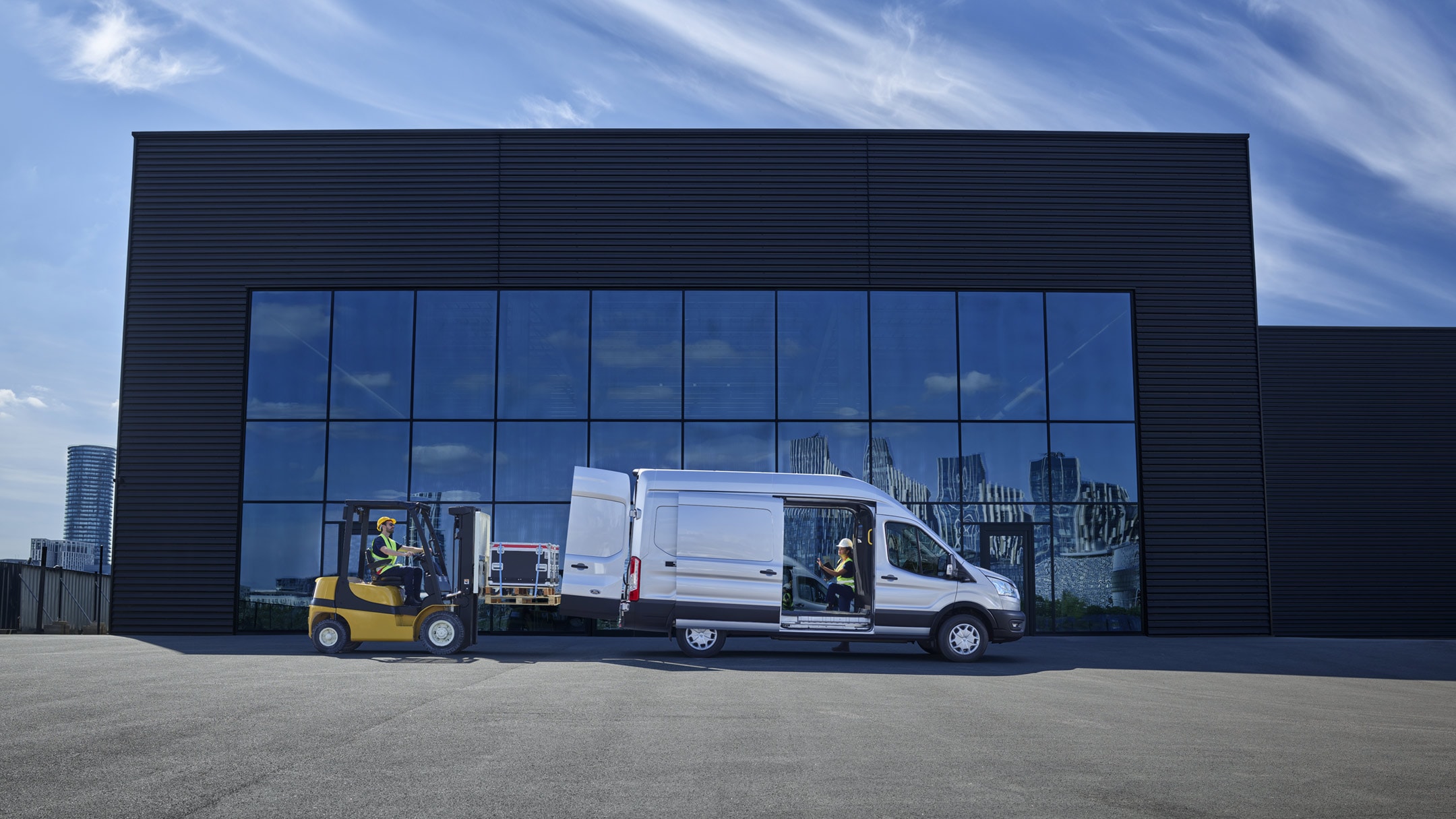 Ford E-Transit Fourgon couleur argent, vue latérale, porte coulissante ouverte et porte arrière en cours de chargement devant un bâtiment moderne