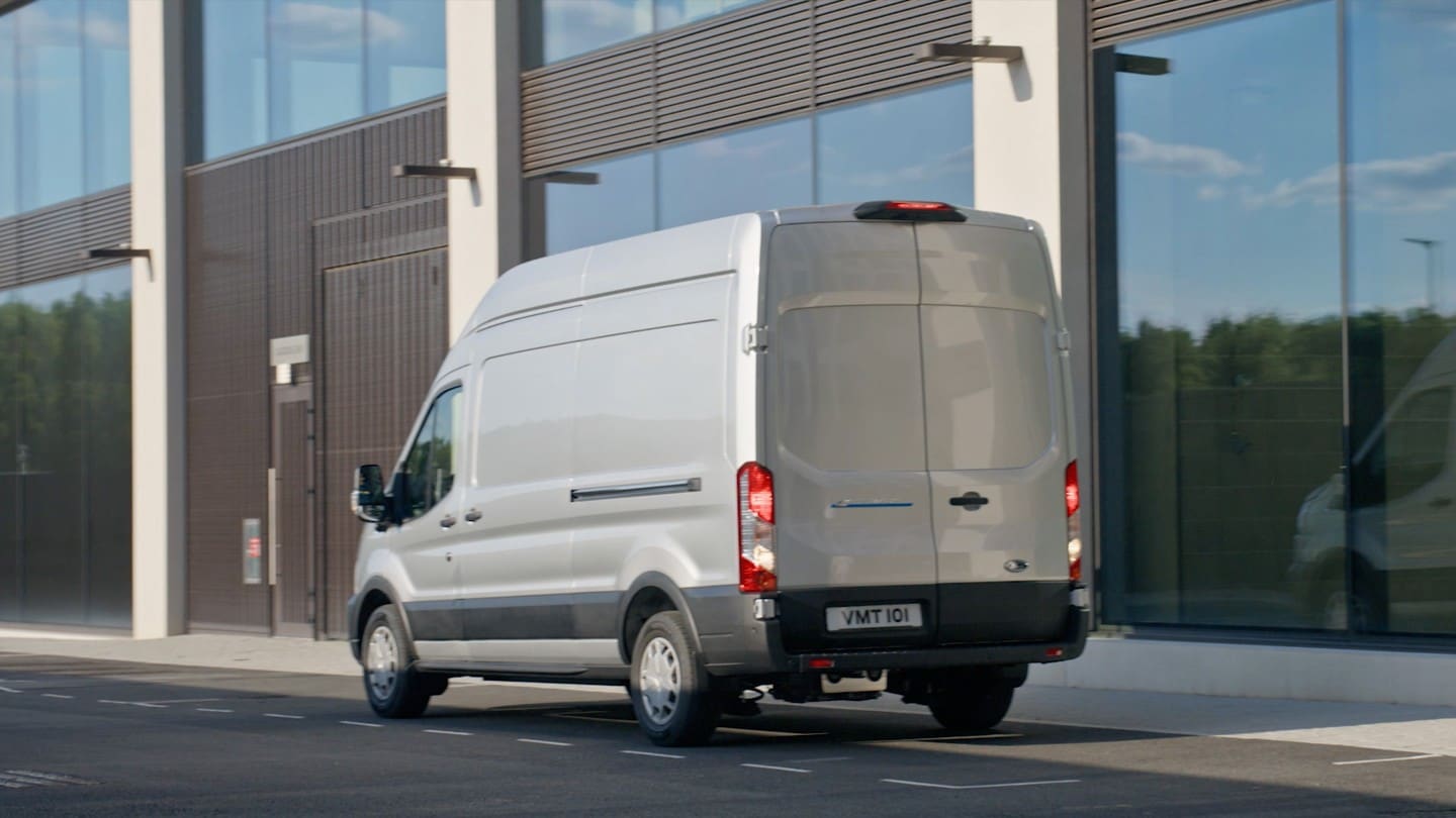 Ford E-Transit, couleur argent, vue arrière aux trois quarts, stationnant devant un bâtiment moderne