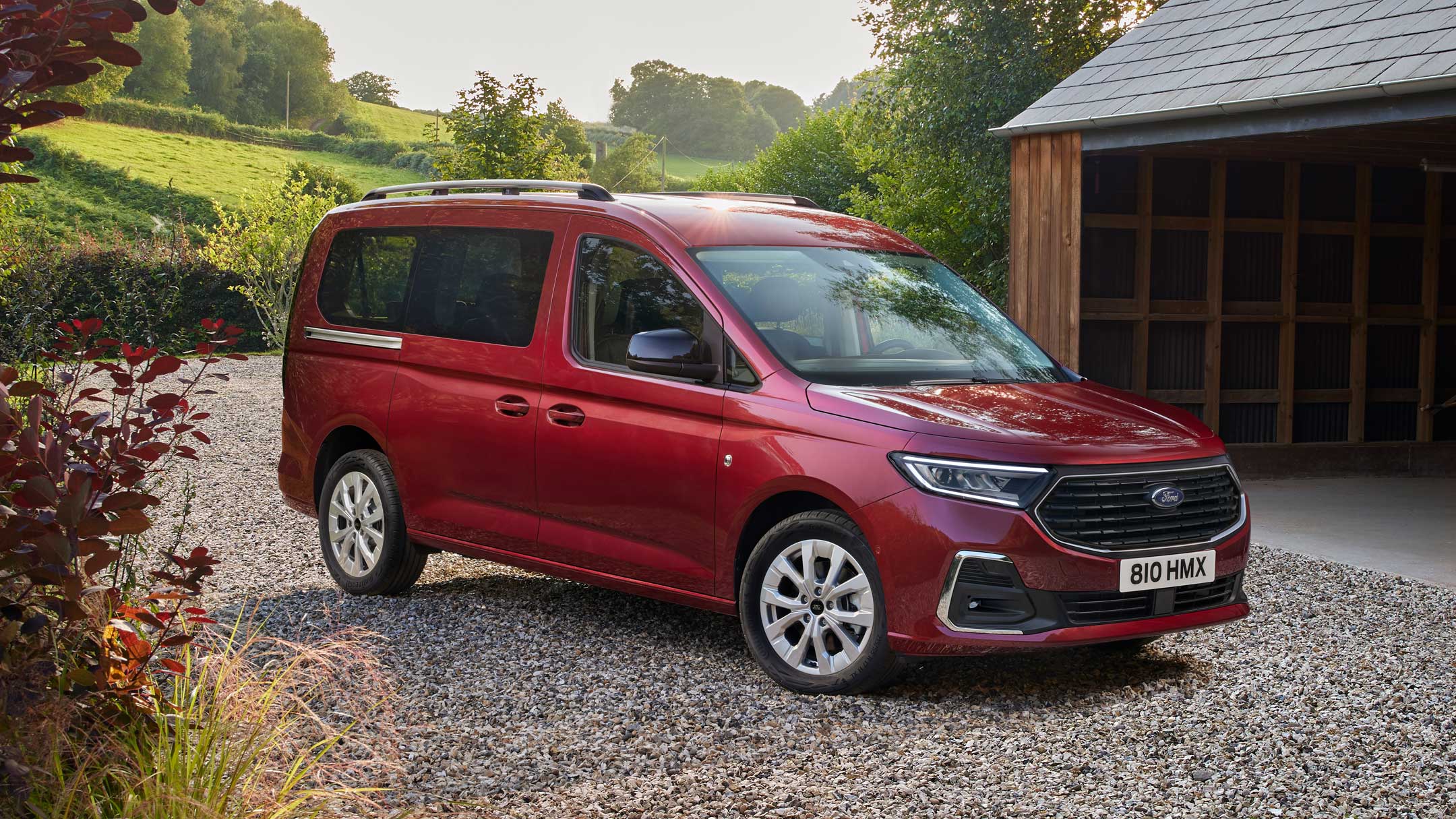 Ford Grand Tourneo Connect couleur rouge, vue de face aux trois quarts, stationnant sur un chemin de gravier, devant une habitation