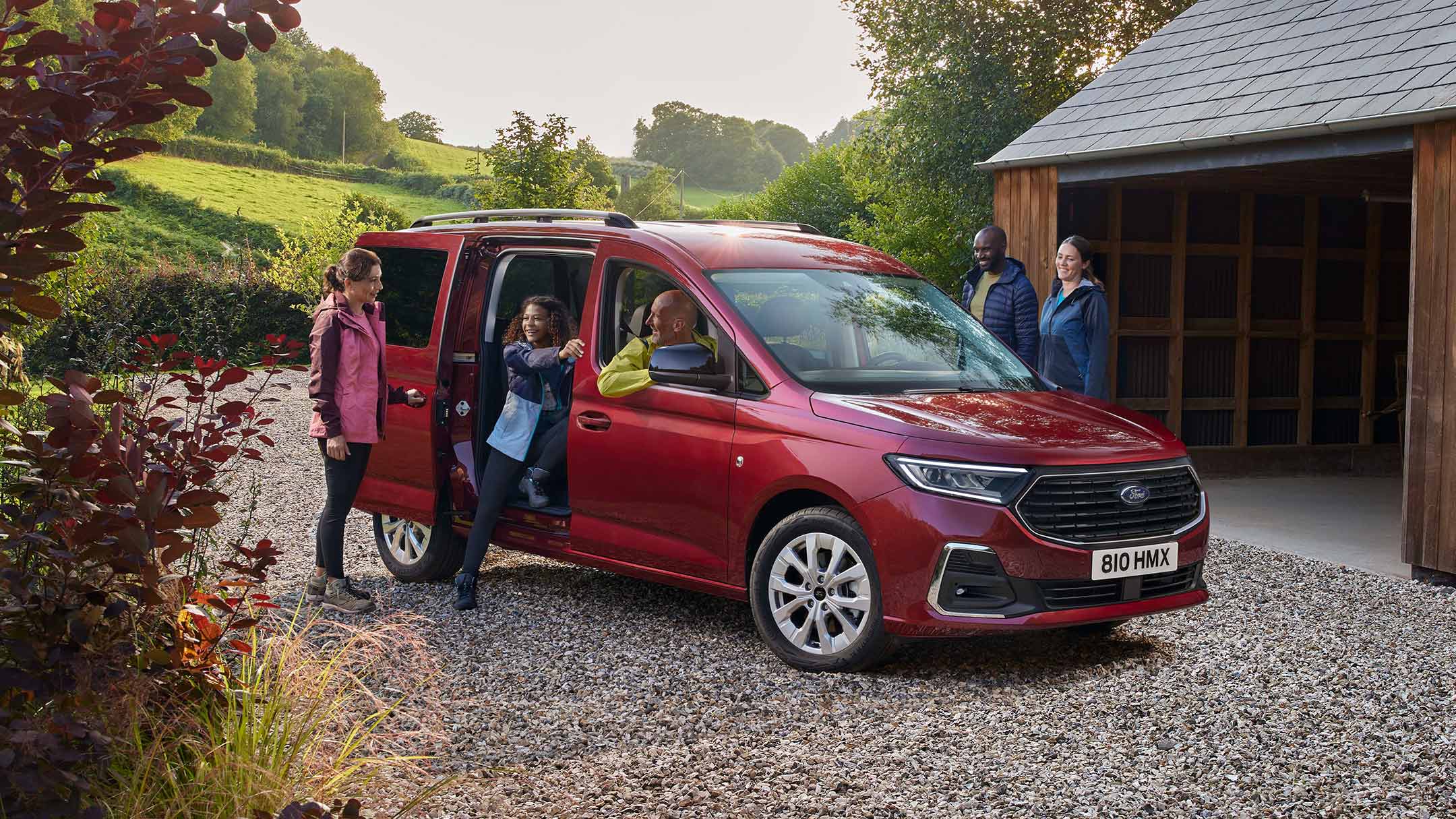 Ford Grand Tourneo Connect couleur rouge, vue de face aux trois quarts, stationnant sur un chemin de gravier, devant une habitation