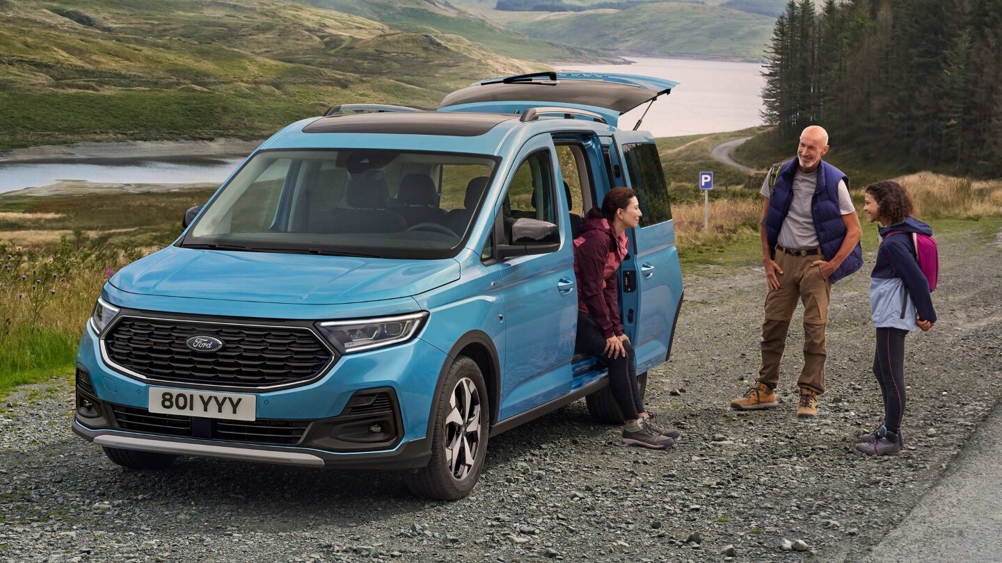 Ford Tourneo Connect Active couleur bleu clair, vue de face aux trois quarts avec porte coulissante ouverte, stationnant sur un chemin de gravier. Femme assise dans le véhicule, son mari et sa fille debout à l’extérieur