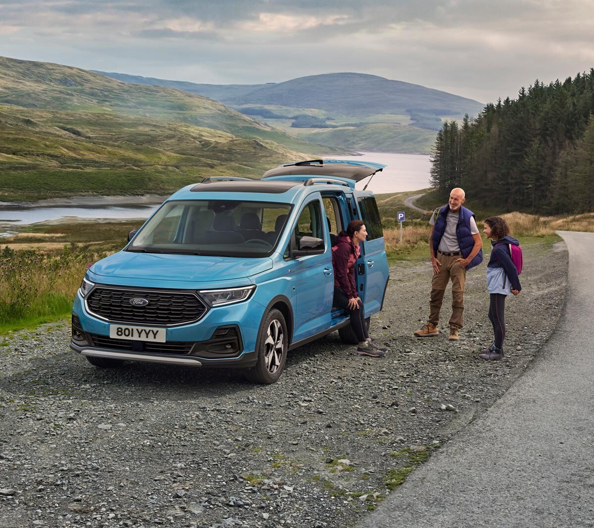 Ford Tourneo Connect Active couleur bleu clair, vue de face aux trois quarts avec porte coulissante ouverte, stationnant sur un chemin de gravier. Femme assise dans le véhicule, son mari et sa fille debout à l’extérieur