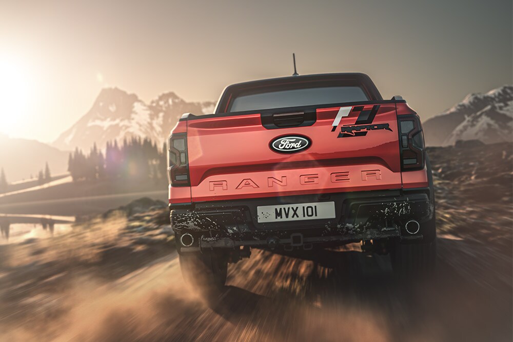 Vue arrière d’un Ford Ranger Raptor rouge circulant sur une piste en terre, avec des montagnes en arrière-plan.