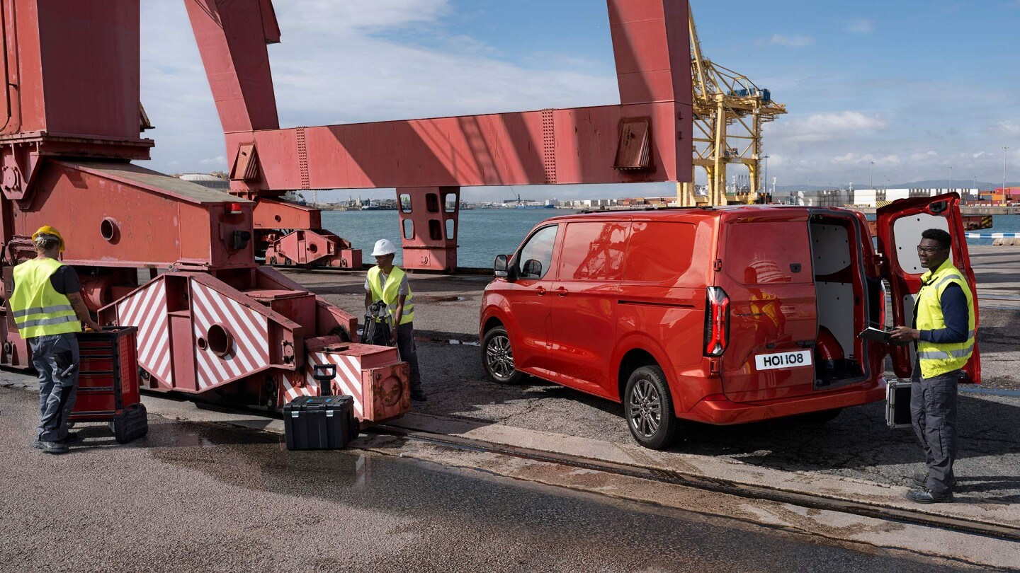 Ford Transit Custom couleur rouge, vue arrière aux trois quarts, en train d’être chargé par des dockers